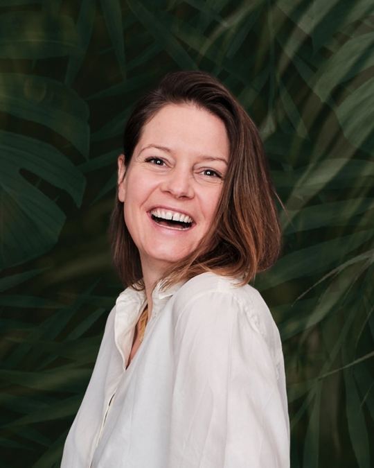 Katharina Baier, Coach und Moderatorin, lachend im weißen Hemd vor grünen Blättern